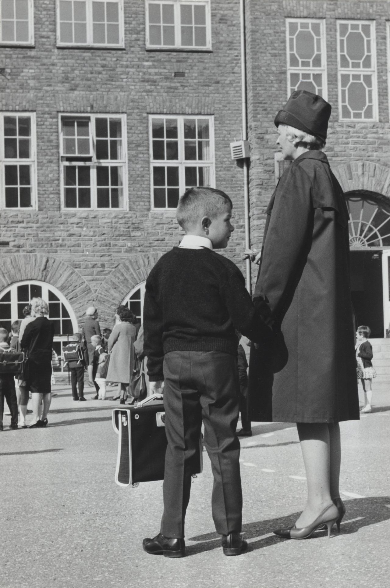 Svart hvitt bilde. Pent kledd ung gutt holder mors hånd i skolegården på første skoledag. I andre hånden har han en skoleveske.