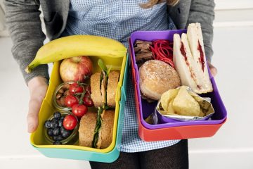 Tett på fanget til en jente i kjole. HUn holder to matpakker i hendene. Ene matpakka inneholder brødskiver, frukt og bær, mens den andre inneholder loff med syltetøy, potetgull og sjokolade.