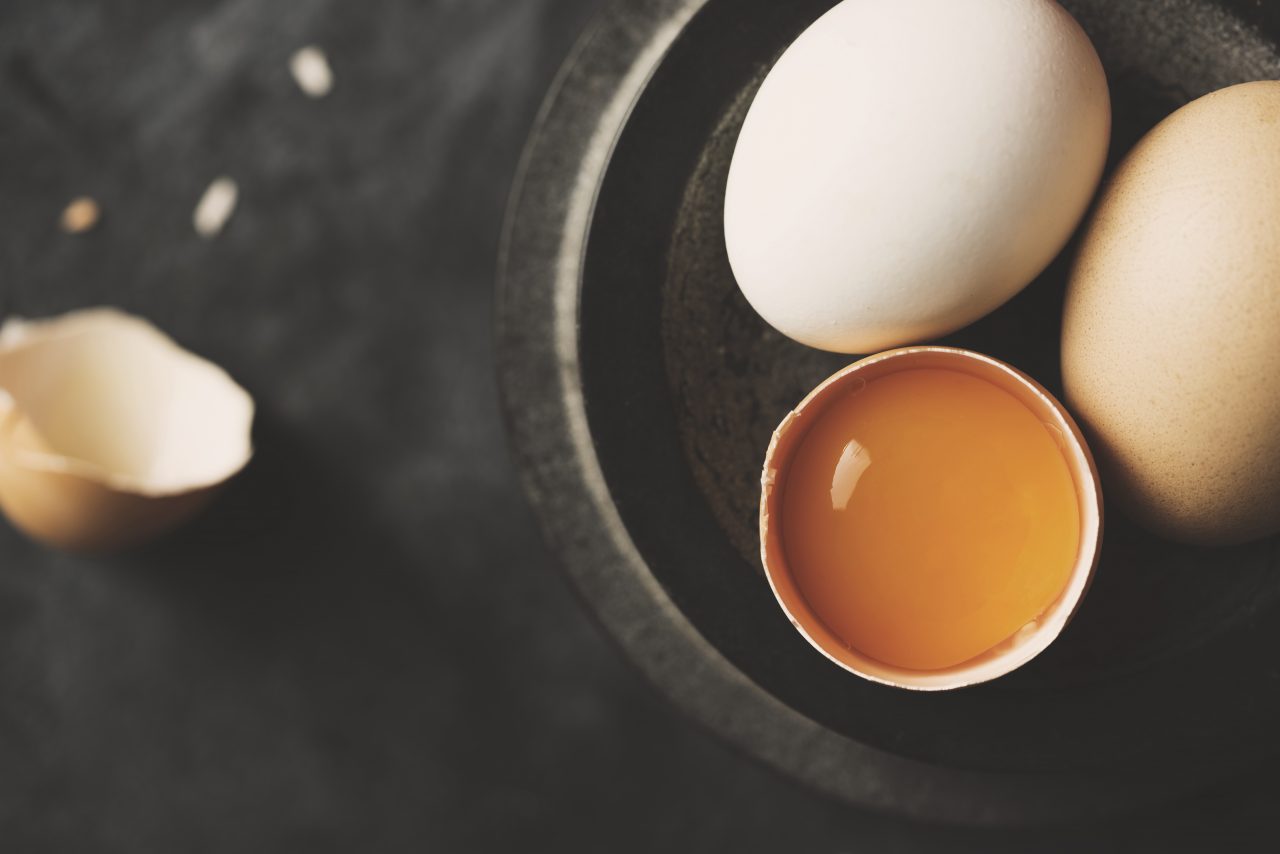 Rått egg i knust skall og to hele egg i en skål, mot en mørk bakgrunn.
