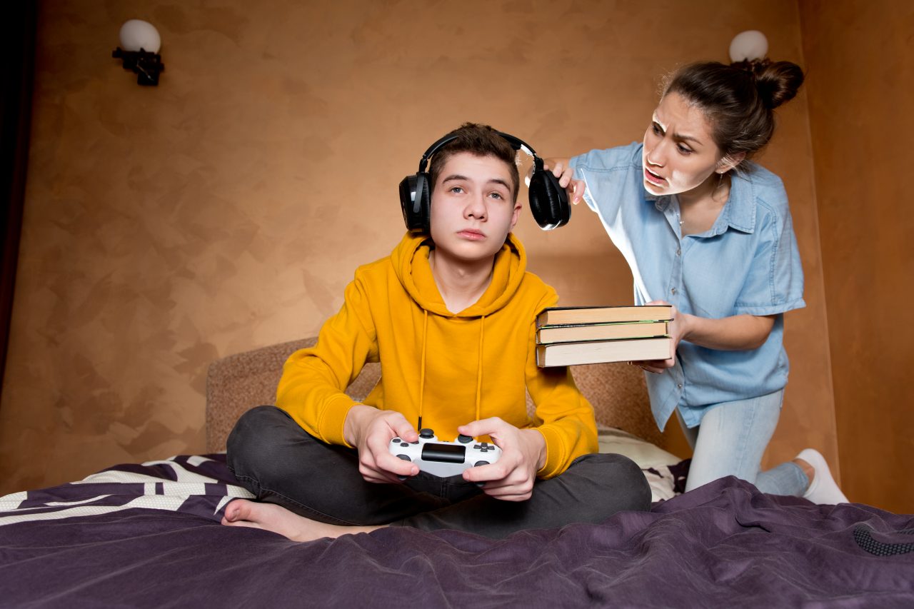 En gutt sitter å gamer i sengen sin. Storesøsteren hans står bak han å prøver å få kontakt med han, fordi han må gjøre leksene sine.