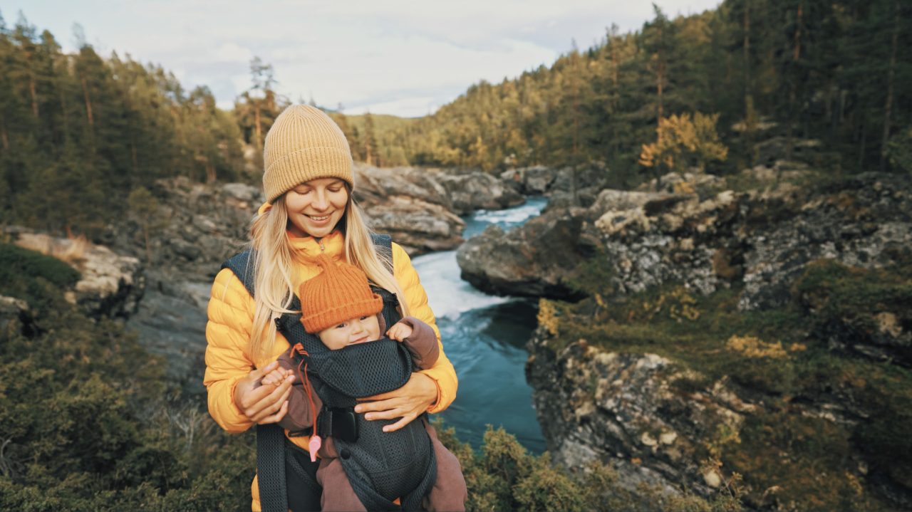 Mor går tur ved en elv på fjellet med baby i bæresele på magen.