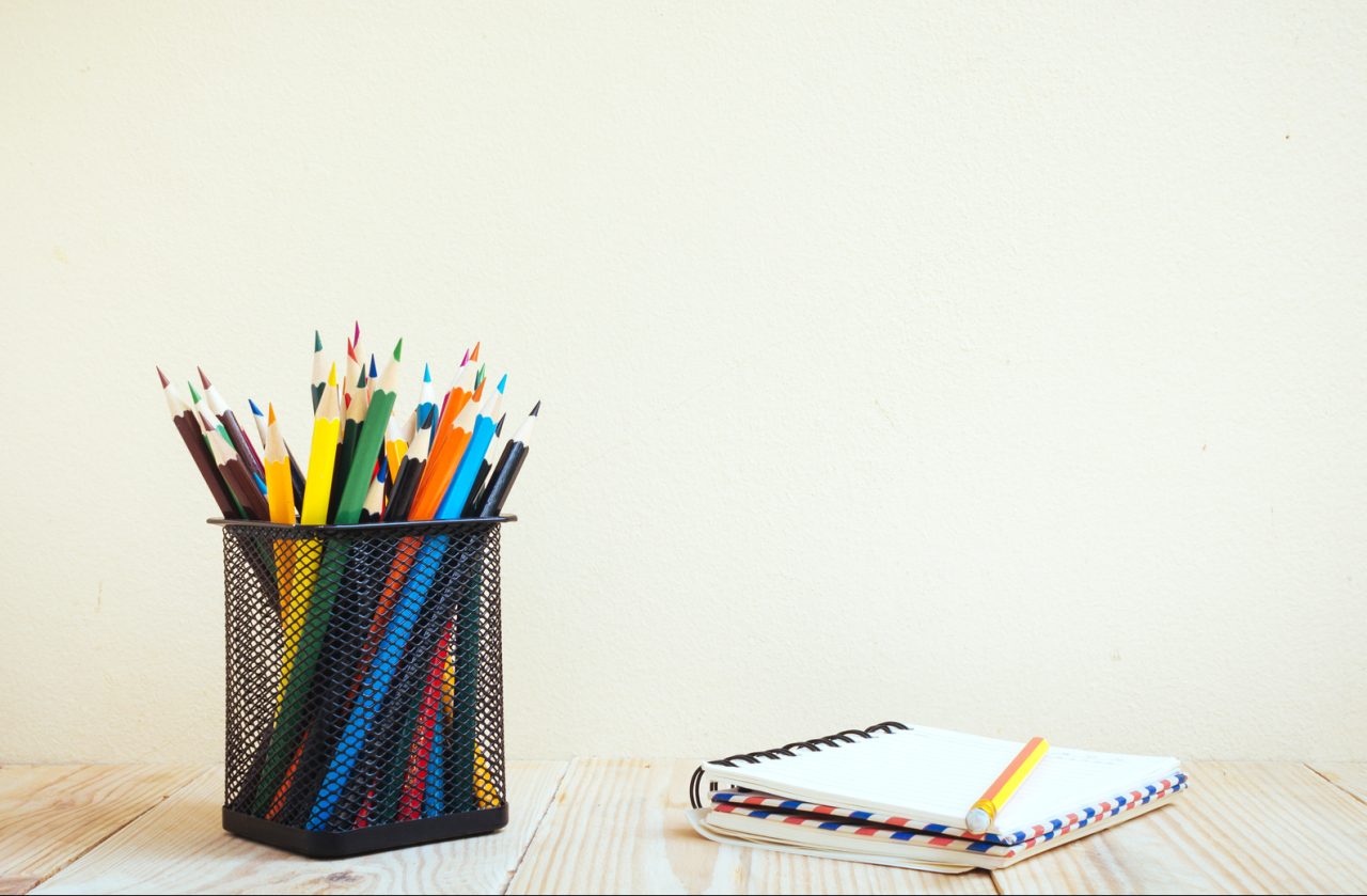 Fargeblyanter i en sort kopp, og en skriveblokk med en blyant liggende oppå.
