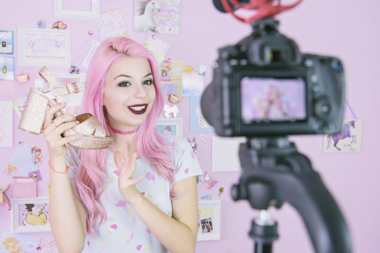 Ung influenser med rosa hår som viser frem en gullsko foran et kamera.
