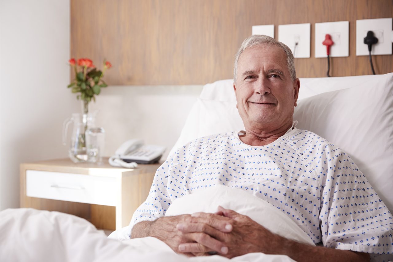 Smilende eldre mann sitter oppreist i en sykeseng.