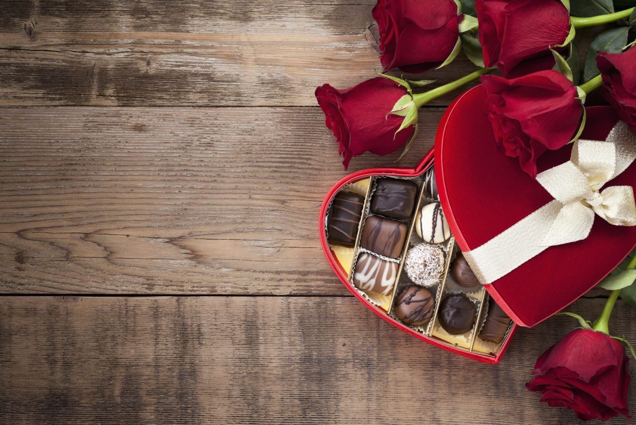 En hjerteformet åpen konfekteske og røde roser liggende på et trebord.