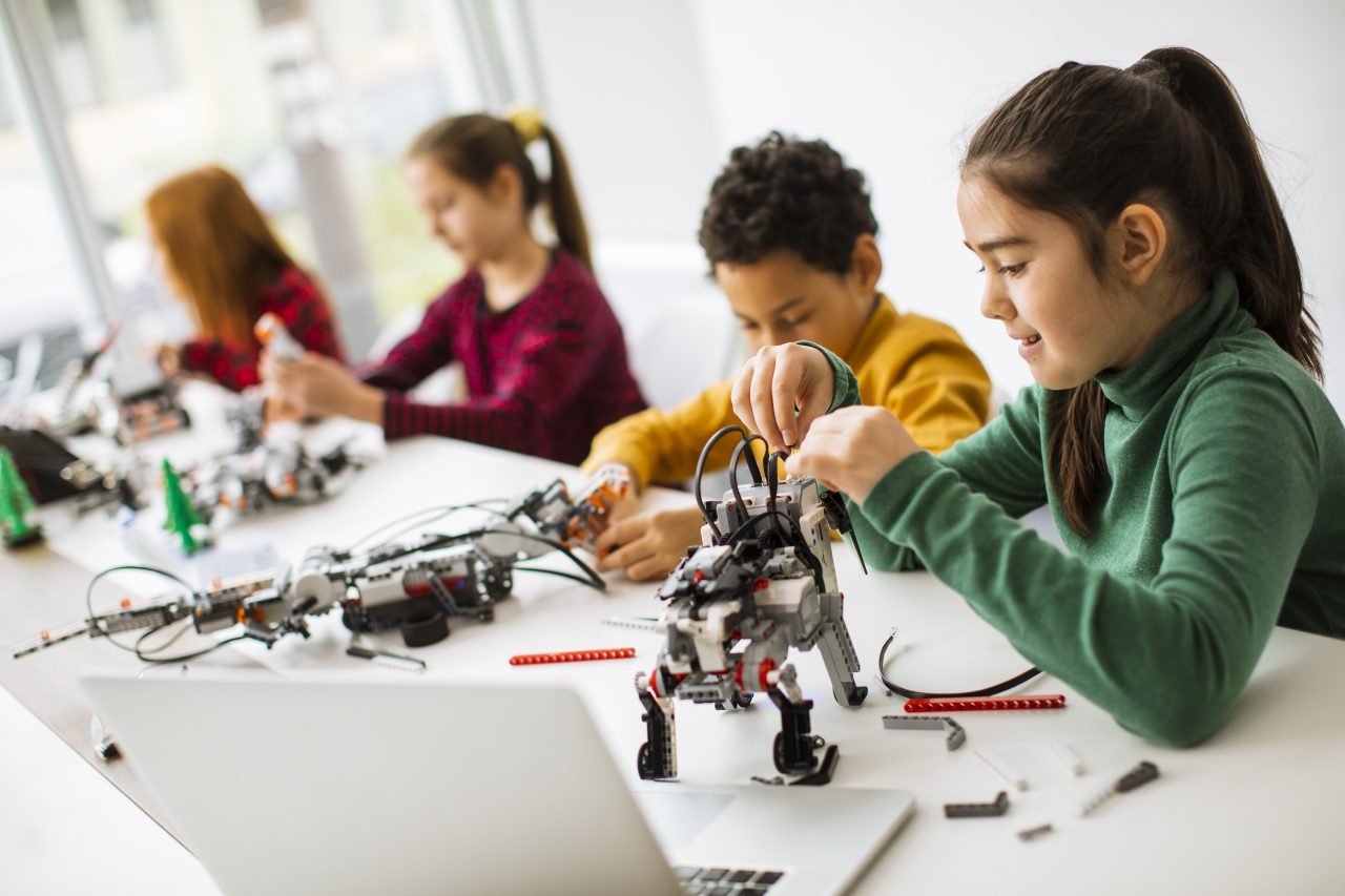 Fire skolerbarn ved et langt bord bygger og programmerer Lego-roboter ved hjelp av digitale instrukser fra en laptop..