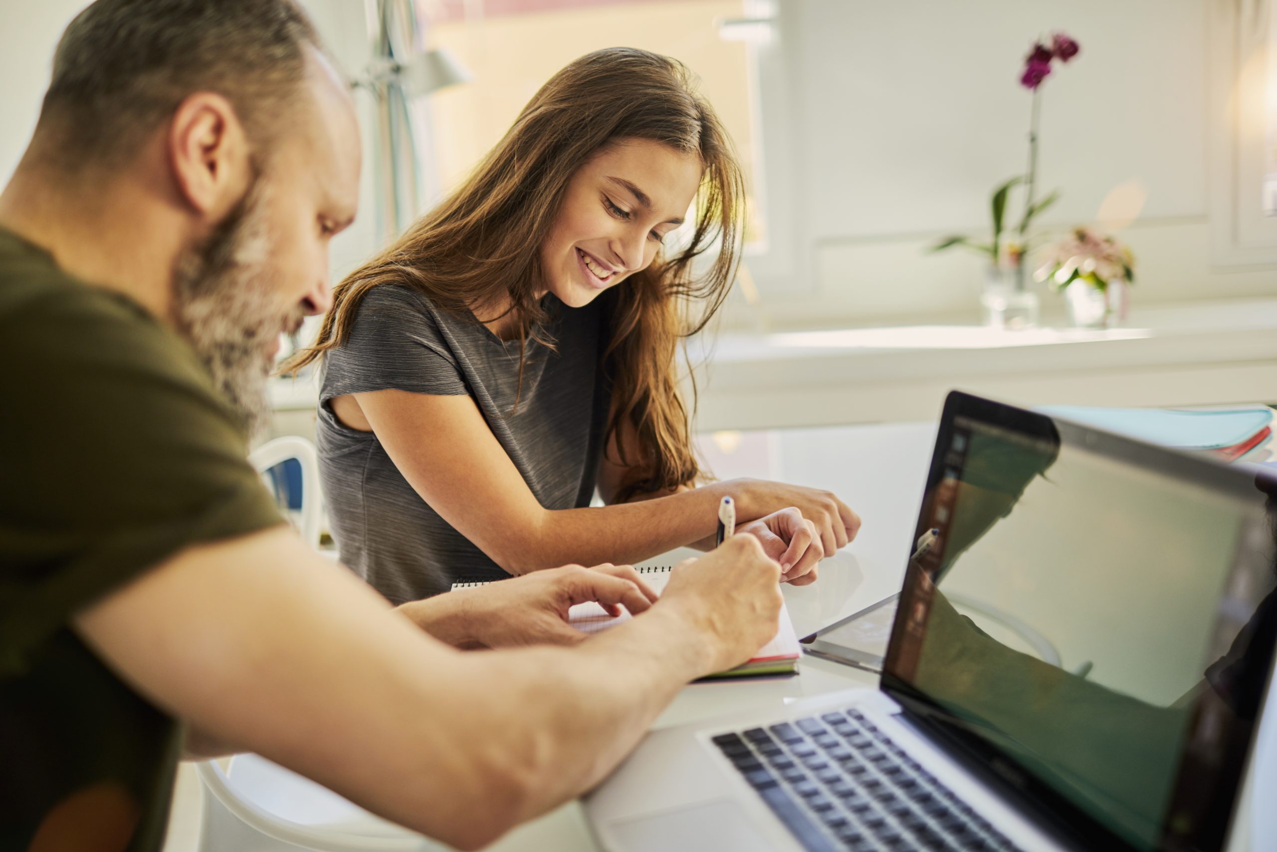 Smilende far og datter rundt spisebord i stuen. Far lener seg bort fra egen laptop for å vise datter noe på blyant og papir.