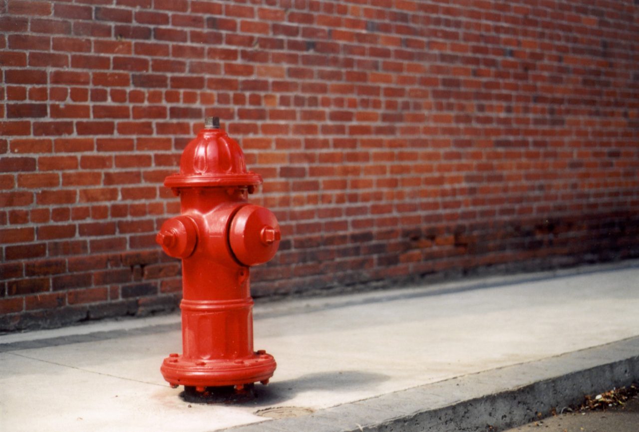 Rød brannhydrant foran en mursteinsvegg.