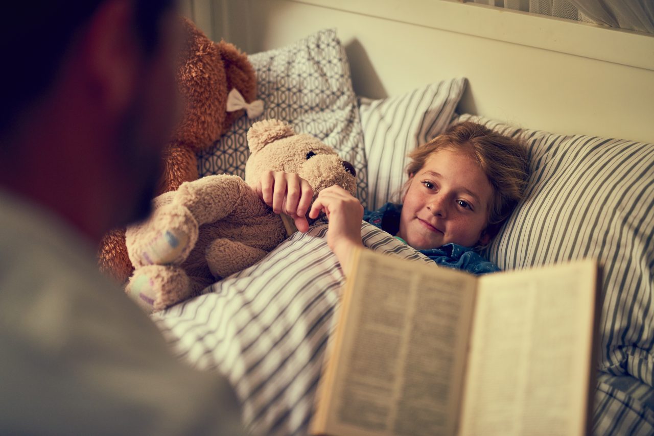 En pappa leser en godnatthistorie for en liten jente som ligger i senga med en bamse.