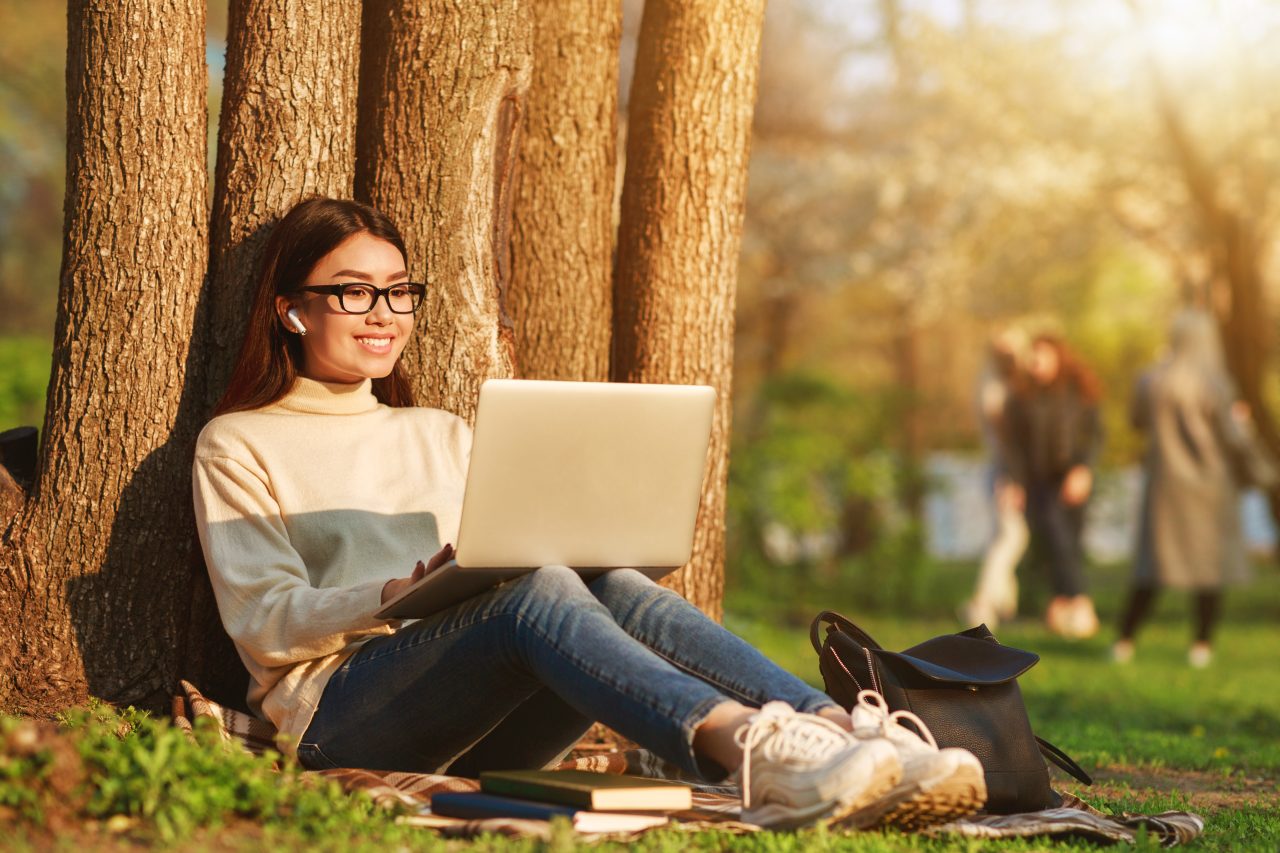 Glad jente sitter lent inntil et tre og jobber på en laptop.