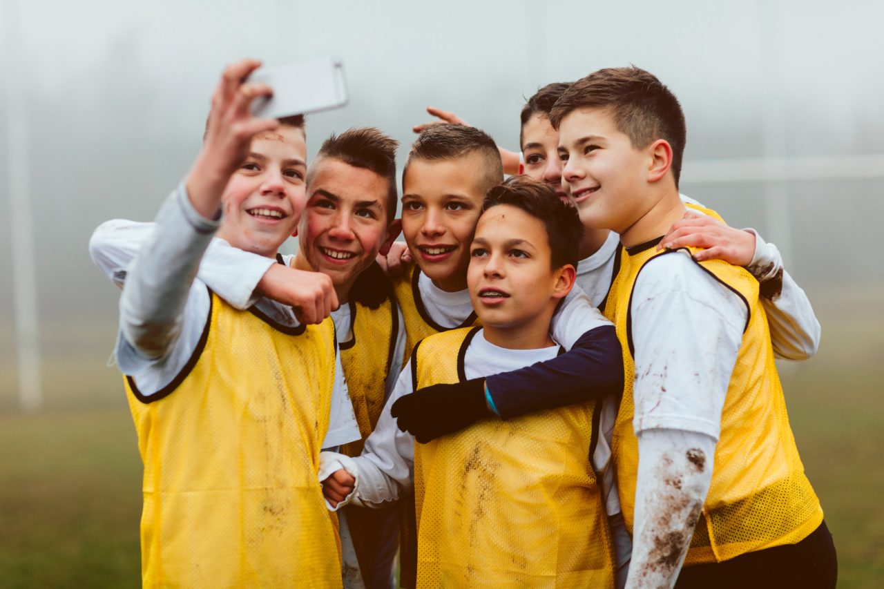 Seks blide gutter med gul vest tar selfie i grått vær på en fotballbane.