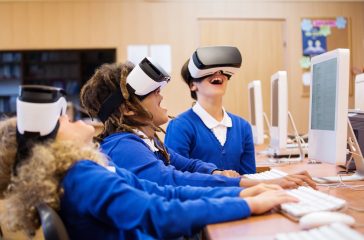 Tre skoleelever sitter ved tre PC-er med blå frakker og VR-briller.