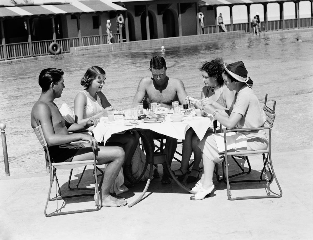 Sort hvitt bilde av fem voksne personer som sitter rundt et bord på stranda og spiser lunsj.