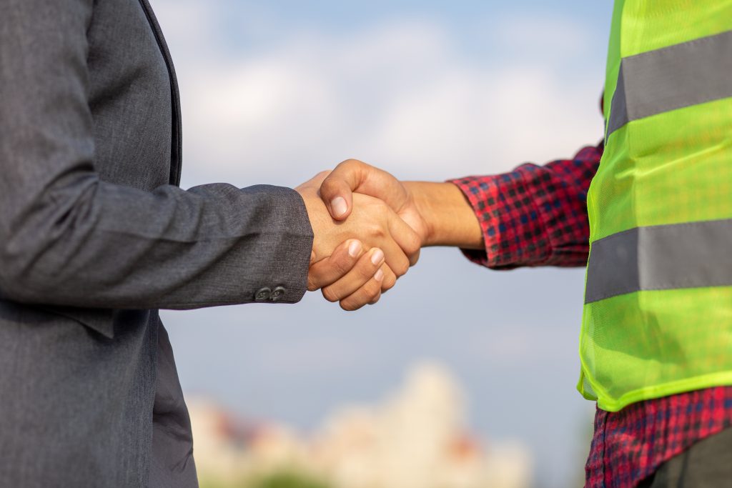 Arbeidsgiver med grå dress og arbeidstaker i gul vest gir hverandre et håndtrykk.