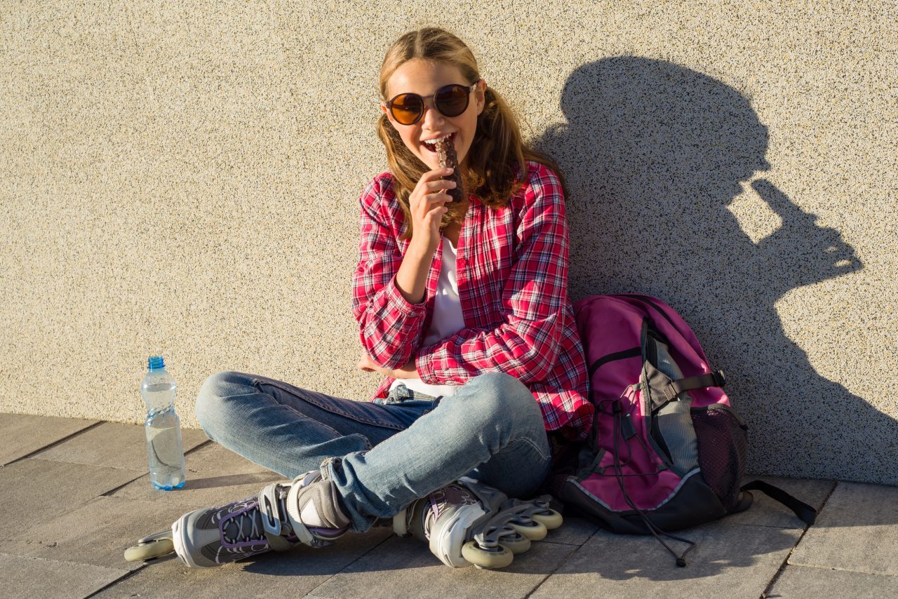 Ung jente med solbriller, skolesekk, rollerblades og vannflaske sitter og spiser sjokolade i solveggen.