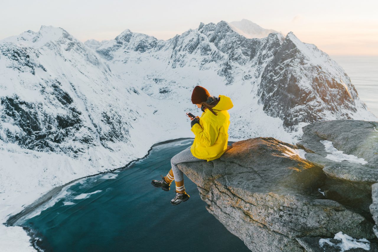 Dame sitter på en en klippe høyt over Lofoten og ser på mobilen