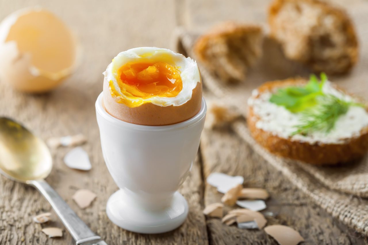 Bløtkokt egg i eggeglass på et bord med eggeskall, skje og brødskiver.