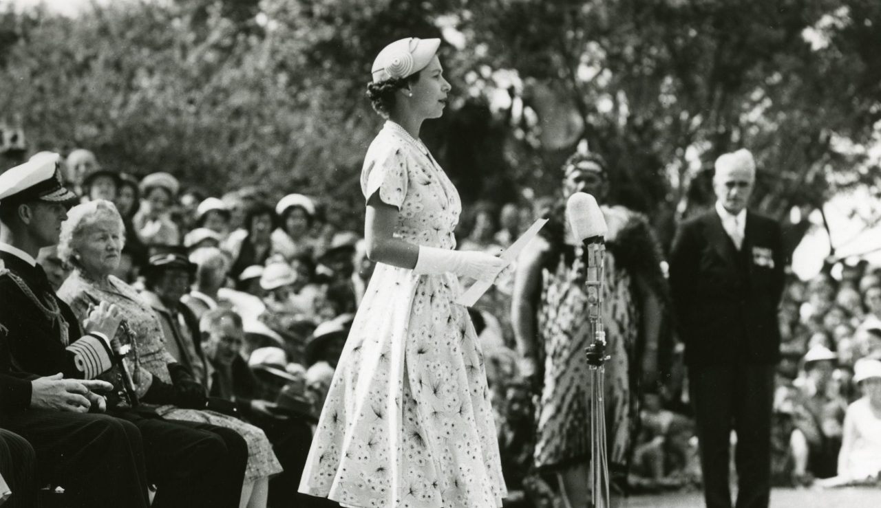 Dronning Elizabeth II står foran mikrofon og taler ikledd lys kjole. Bak henne sitter en forsamling med mennesker.