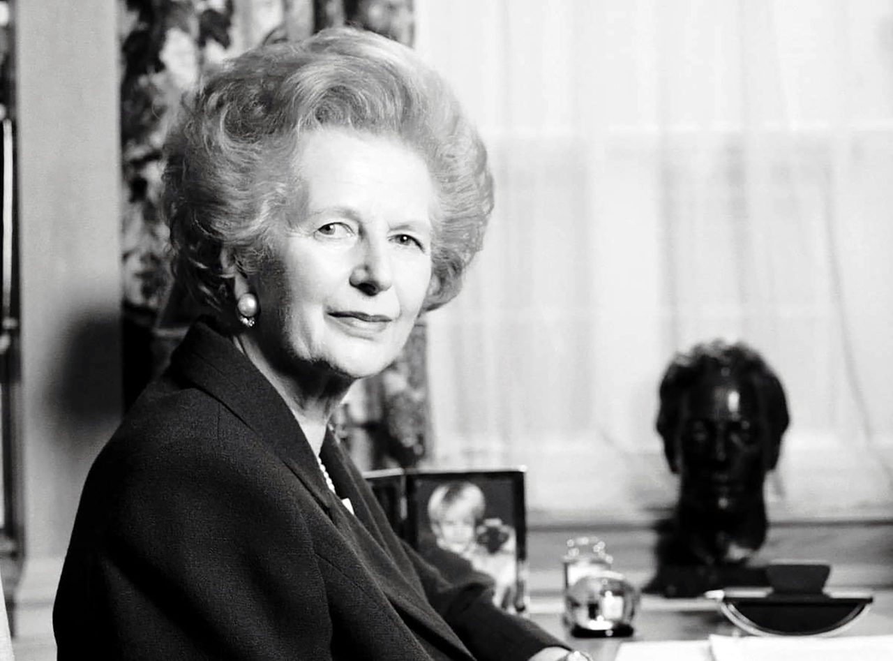 Sort/hvitt-bilde av Margaret Thatcher sittende sidelengs foran skrivepult med ansiktet vendt mot kamera.