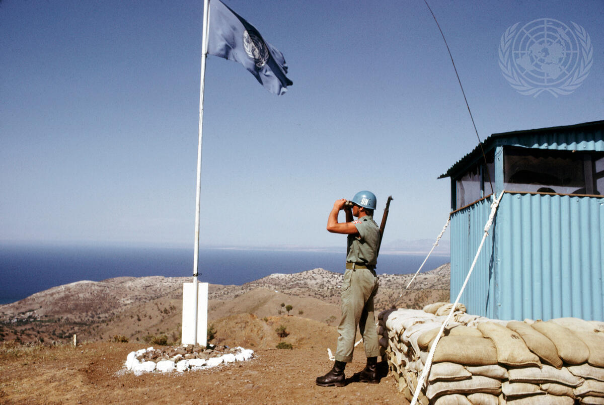 FN soldat står på en høyde og speider utover med en kikkert.