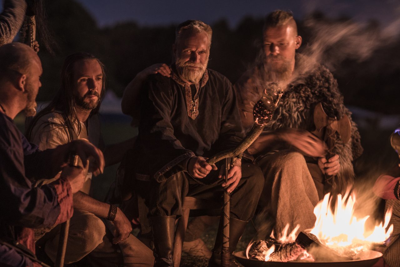 En gjeng med vikinger sitter rundt et leirbål og har det hyggelig sammen.