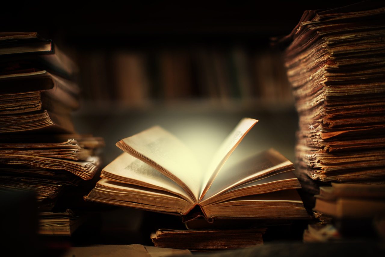 Magisk bok ligger åpen på et skrivebord med stabler av bøker på hver side, og sidene i boka lyser.