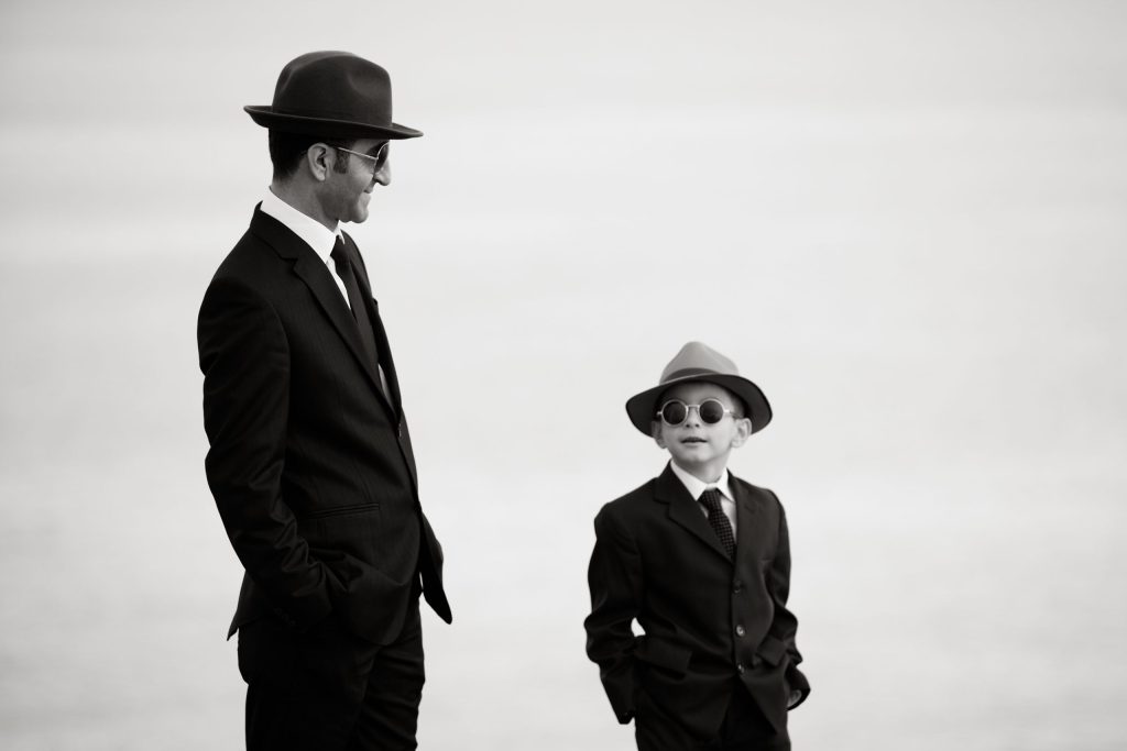 Sorthvitt bilde av en far og ung sønn der sønnen er kledd som faren i dress og hatt.