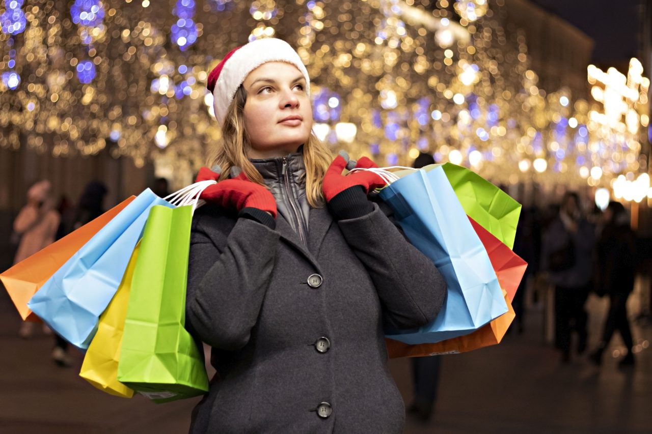 Dame med rød nisselua holder mange handleposer i hendene sine foran et opplyst shoppingsenter.