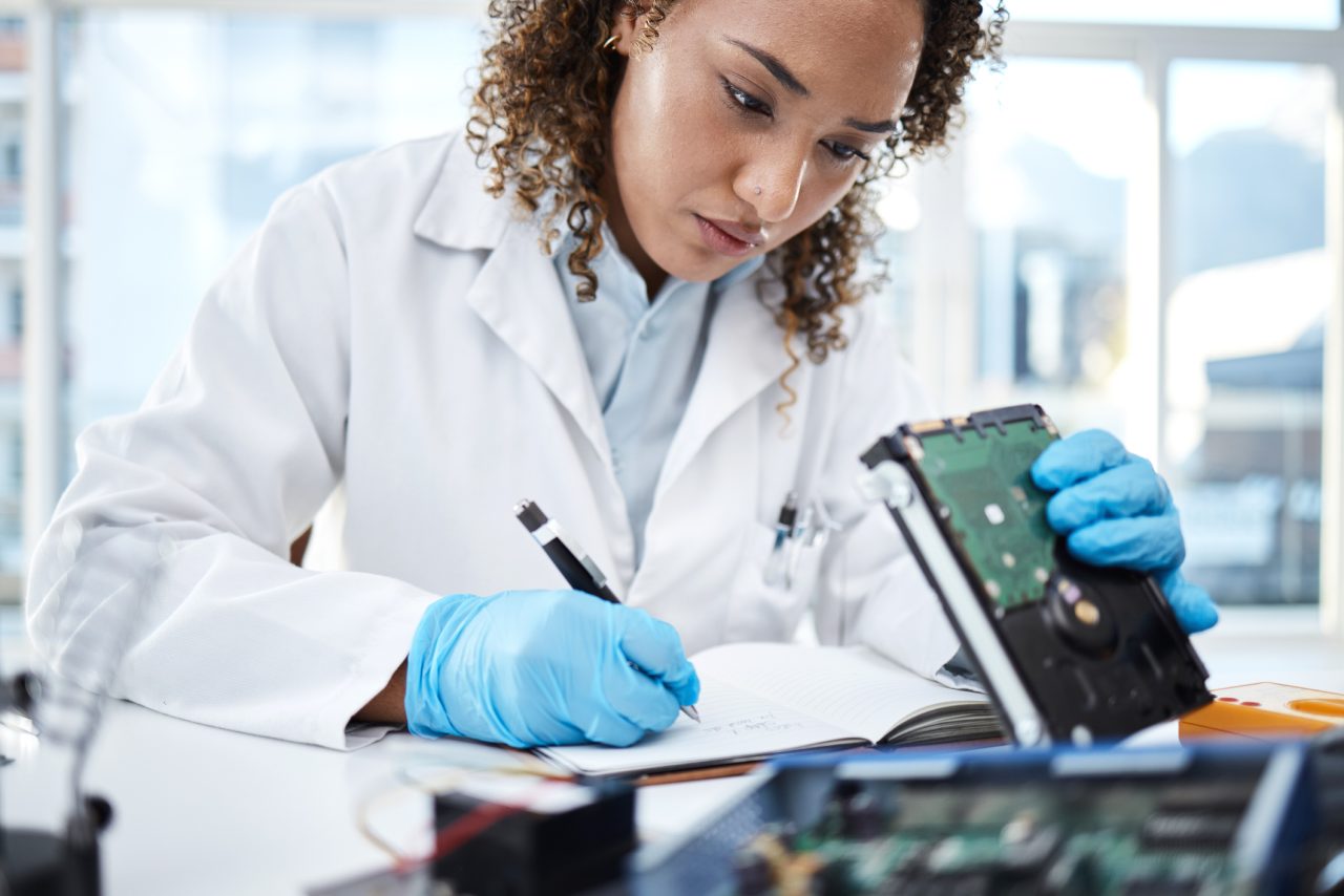 Kvinnelig tekniker i hvit labfrakk som jobber med en harddisk og noterer på et ark