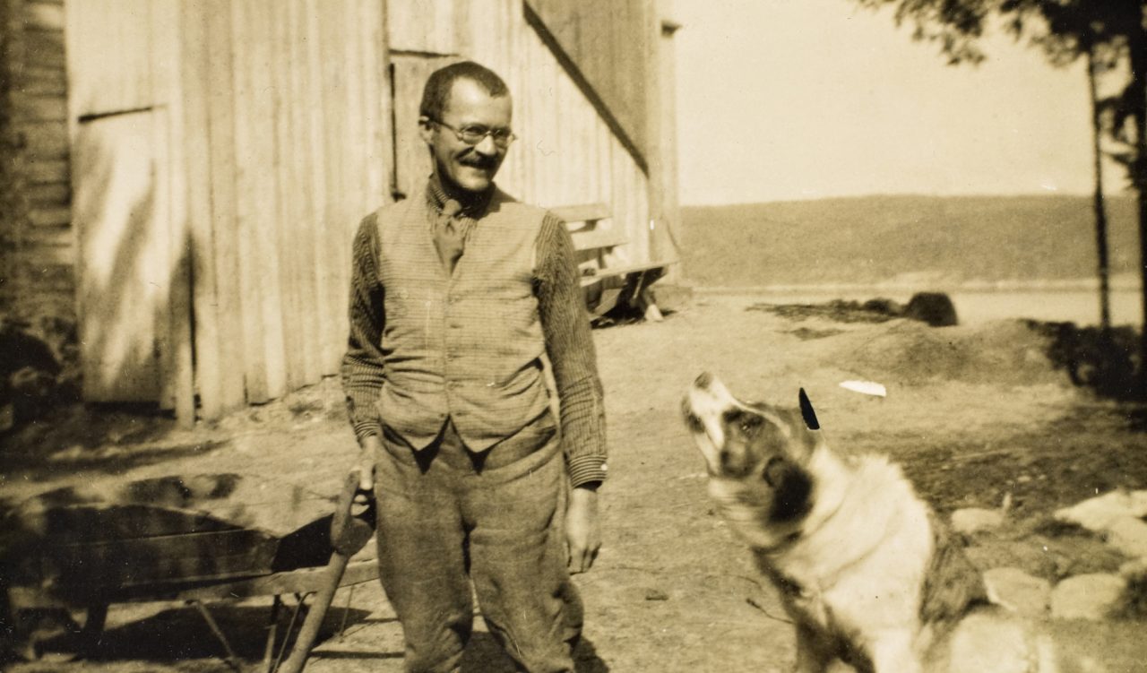 Arnulf Øverland med en spade i hånden og en hund ved siden av seg, stående foran en låve.