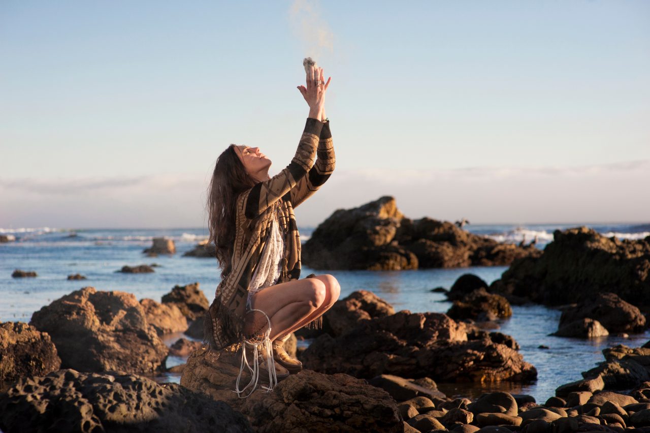En kvinne sitter i fjæra og tilber naturen, hun brenner røkelse