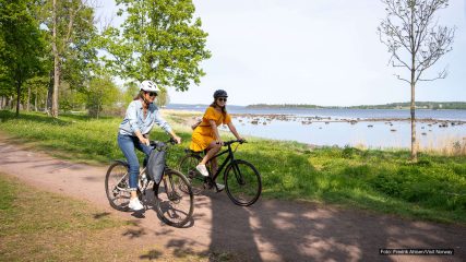 To personer sykler på en grussti langs kysten, med grønt landskap, trær og sjø i bakgrunnen.