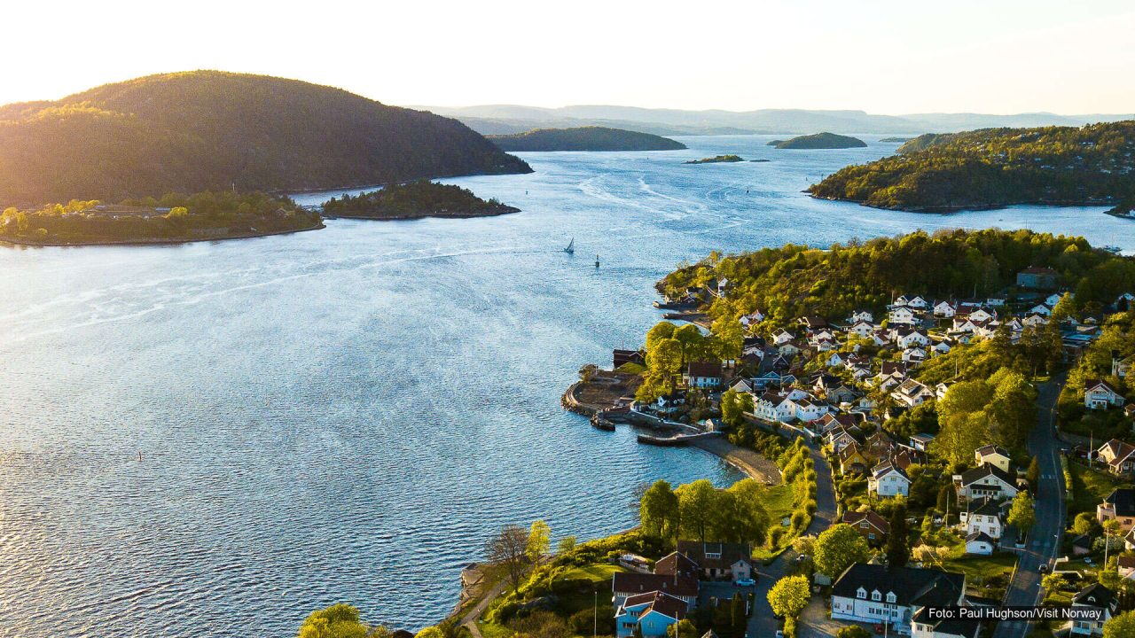Dronebilde av Drøbak med tett bebyggelse langs fjorden, grønne åser og små øyer i bakgrunnen.