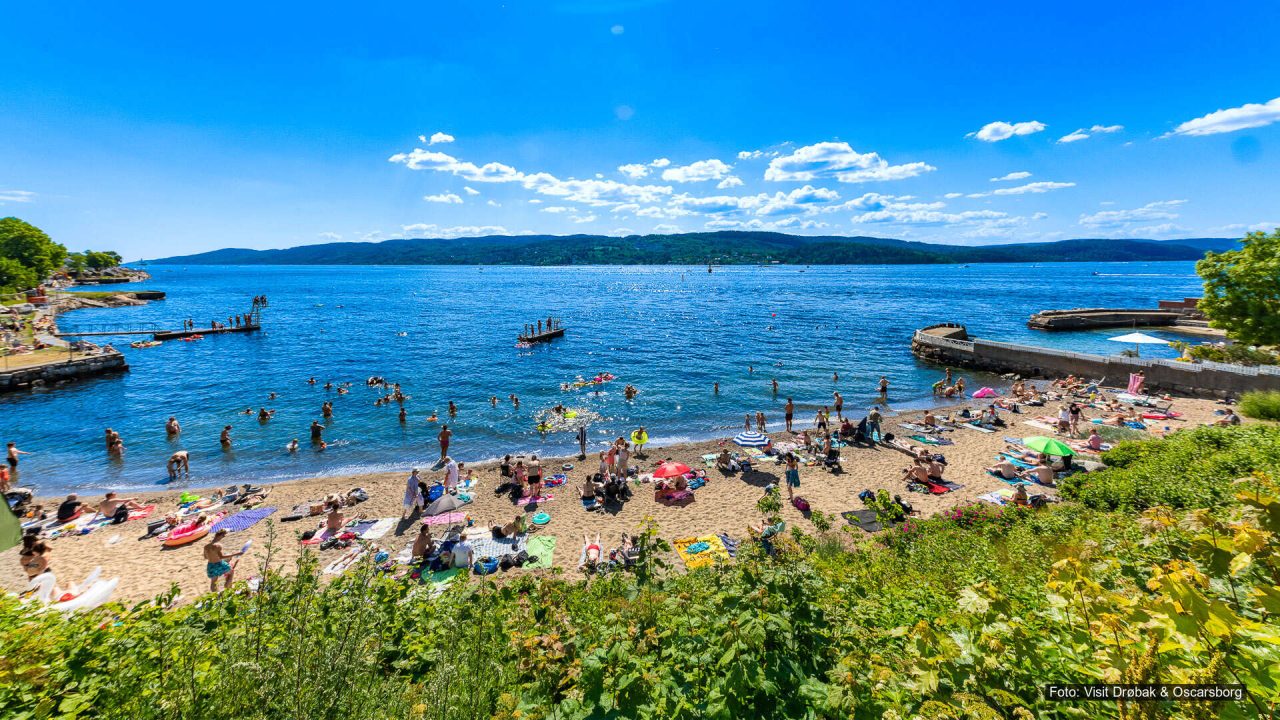 Mange mennesker bader og soler seg på sandstrand ved fjorden på en solrik sommerdag.