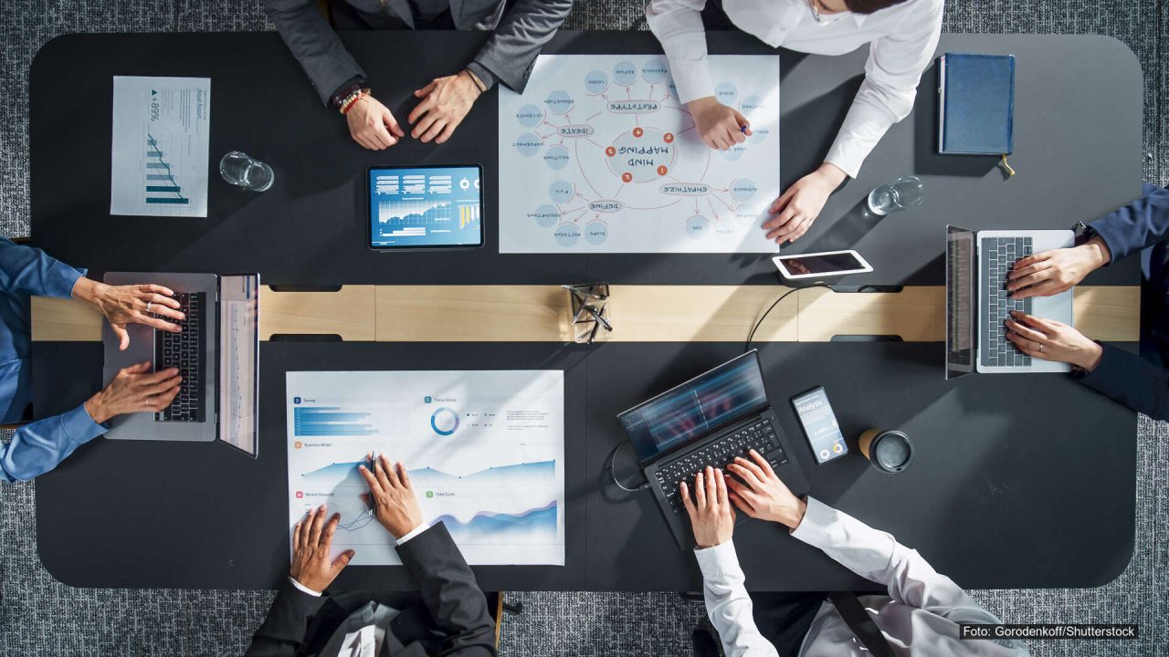 Personer rundt et møtebord jobber med diagrammer, datamaskiner og nettbrett sett ovenfra.