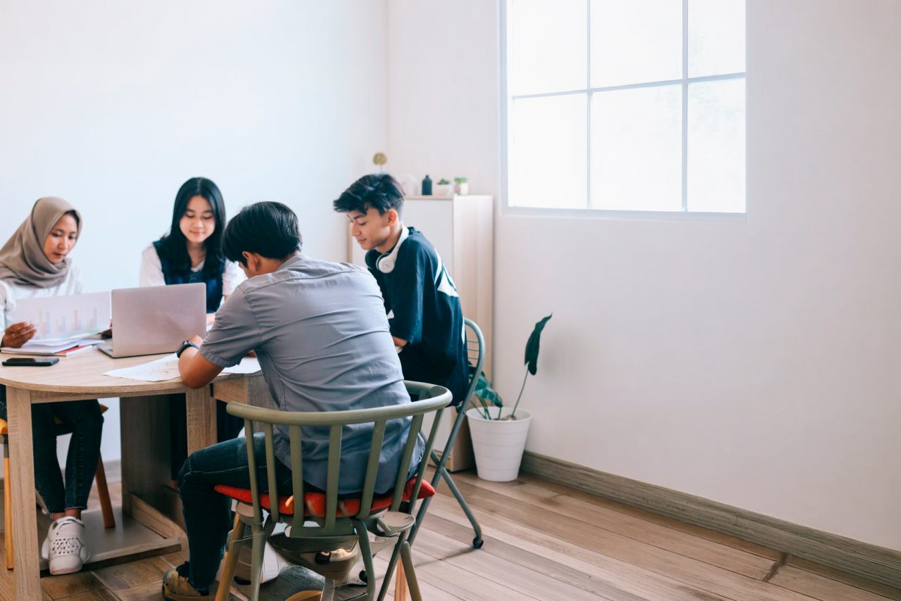 Studenter som sitter sammen ved et bord med bøker og bærbare datamaskiner, gruppearbeid for å diskutere et prosjekt i klasserommet